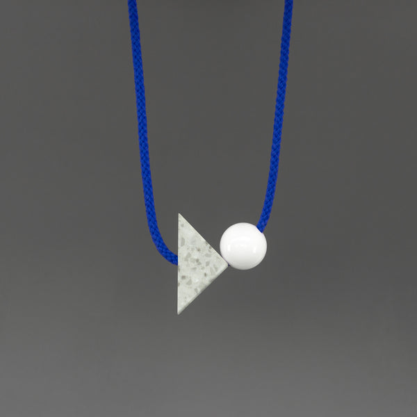 The Brandt necklace is a bright and bold statement. Stepping away from our signature black cord, Brandt's bright blue cord offers something different and looks great against a variety of tops.  An off-white triangle of corian with white and grey speckles (70mm x 50mm) and a white resin ball (30mm). 
